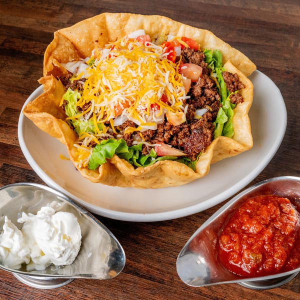 A taco salad in a crispy tortilla bowl with ground beef, lettuce, tomatoes, onions, and shredded cheese, served with salsa and sour cream.