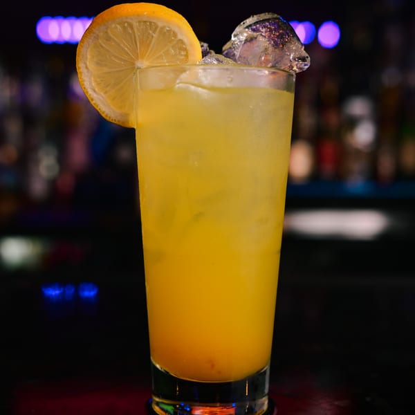 A tall glass of yellow cocktail with ice and a lemon slice, against a blurred bar background.