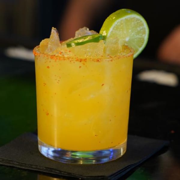 A vibrant yellow cocktail with ice, a lime slice, and jalapeño, garnished with chili salt.