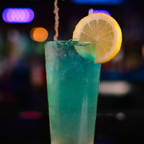 Blue cocktail with a lemon slice, liquid splashing, and blurred colorful lights in the background.