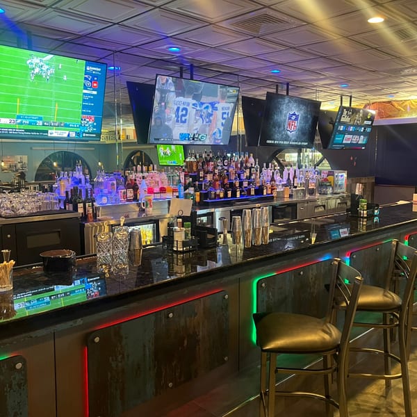A well-stocked bar at Foodys Tampa with multiple TVs showing sports, illuminated by colorful LED lights.