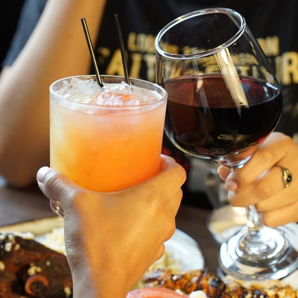 Two people clinking glasses, one with a red wine, the other with an orange cocktail.