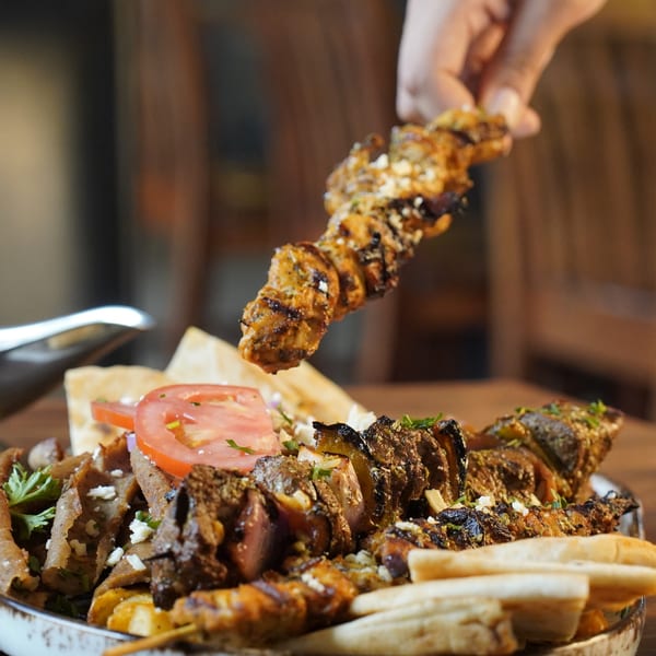 A hand holds a grilled chicken skewer over a platter of Foodys Tampa's gyros, tomatoes, and pita.