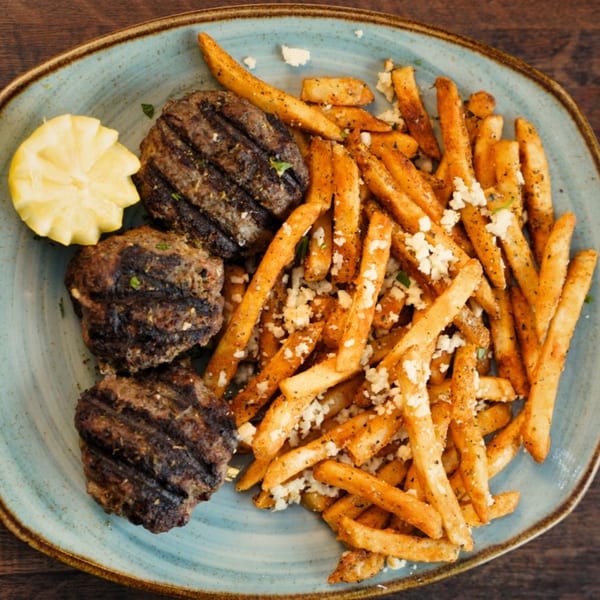 Three grilled patties with a side of seasoned fries topped with feta cheese and a lemon wedge.